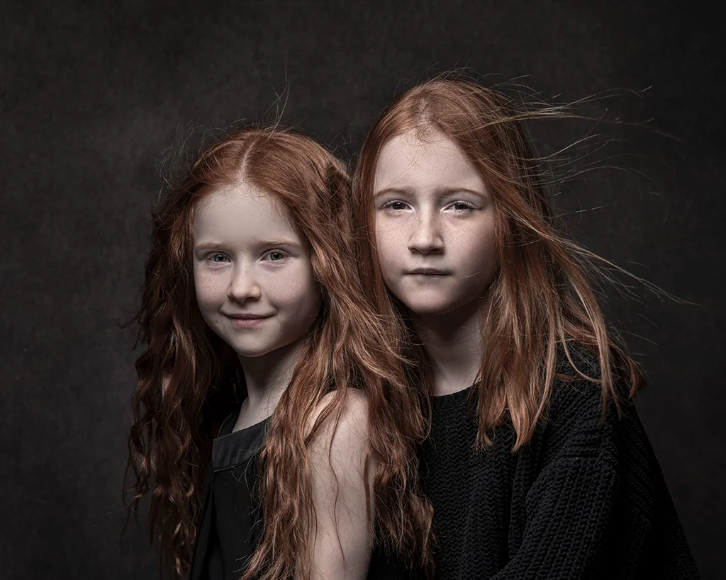 Two girls with red windswept hair wearing dark clothing on a dark background.