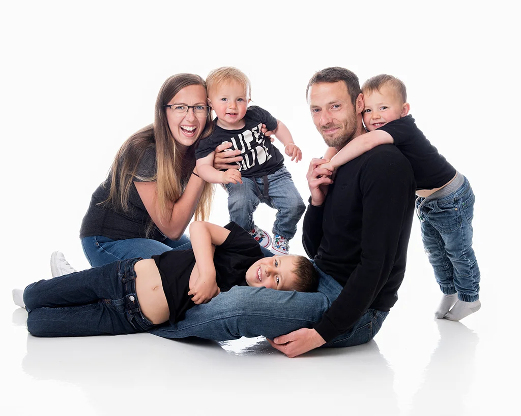 A family of mum, dad and three young boys, all wearing black tops and blue jeans, happily play in a very white room