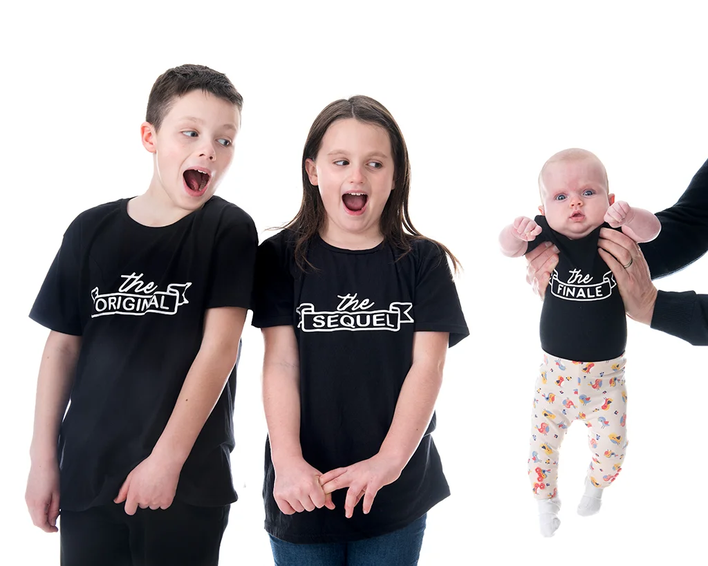 Boy wearing black t-shirt with The Original on it with sister in the same but with The Sequel standing to his left. New baby in black t-shirt with The Finale on it held from right hand side by parent.