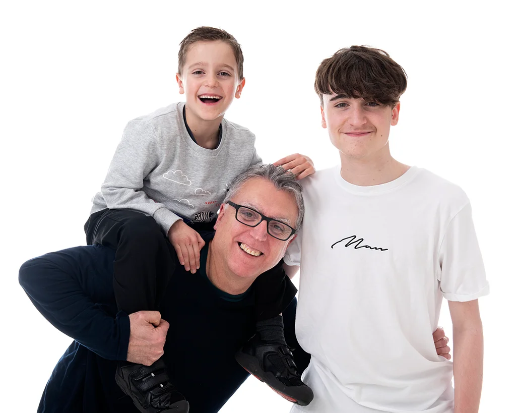 A dad in black top with his two sons, the younger sitting on his shoulders and the tall older boy at his side in a very white room