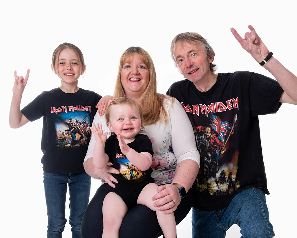 Nan in pale top sitting with her husband and two granddaughters who are all wearing black Iron Maiden t-shirts and make to sign of the beast in a completely white room