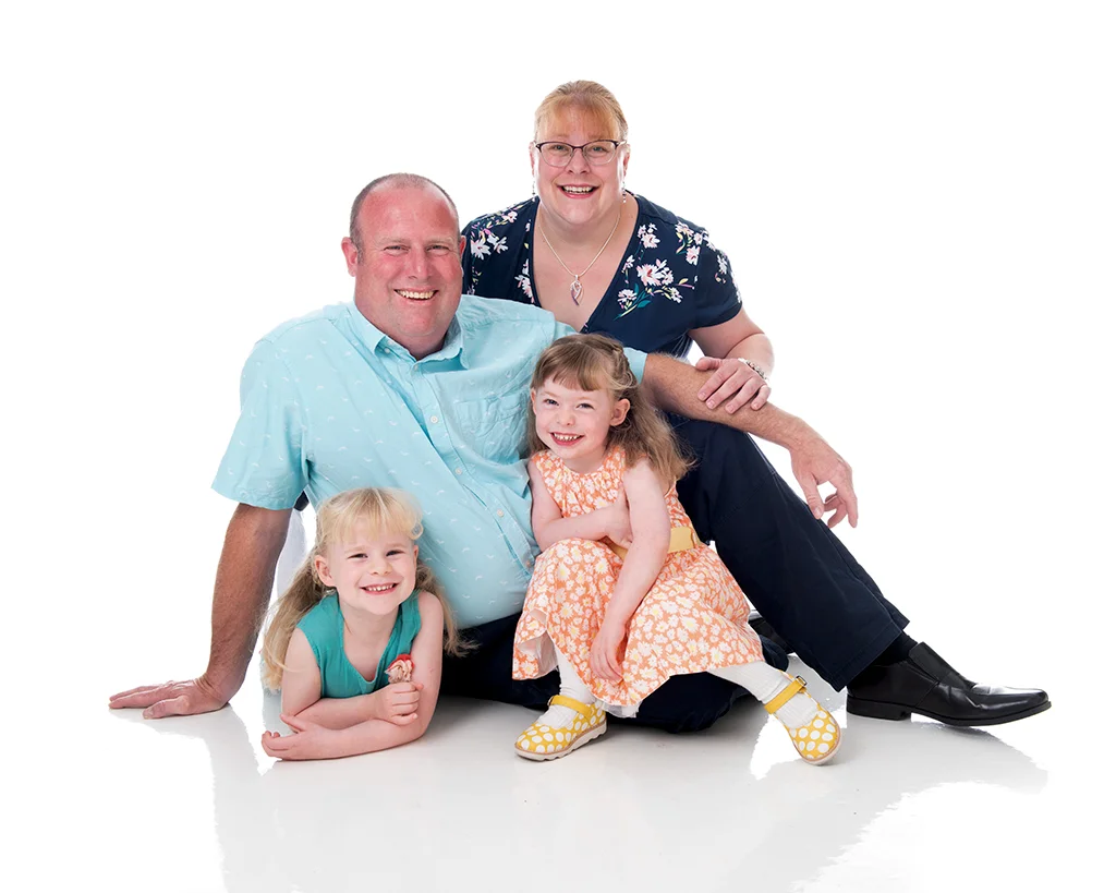 Mum in blue dress behind her husband in a light blue shirt sitting on the floor of a white room with their two daughters