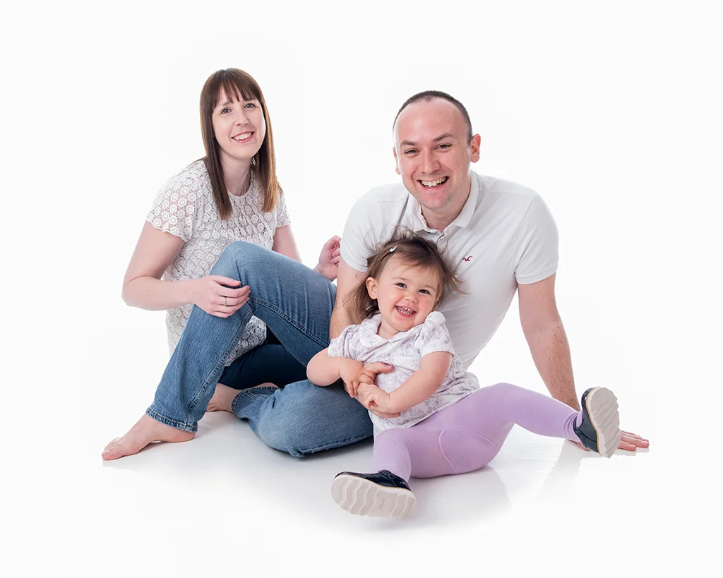 Mum and dad sitting on the floor with their two year old daughter laughing in a completely white room
