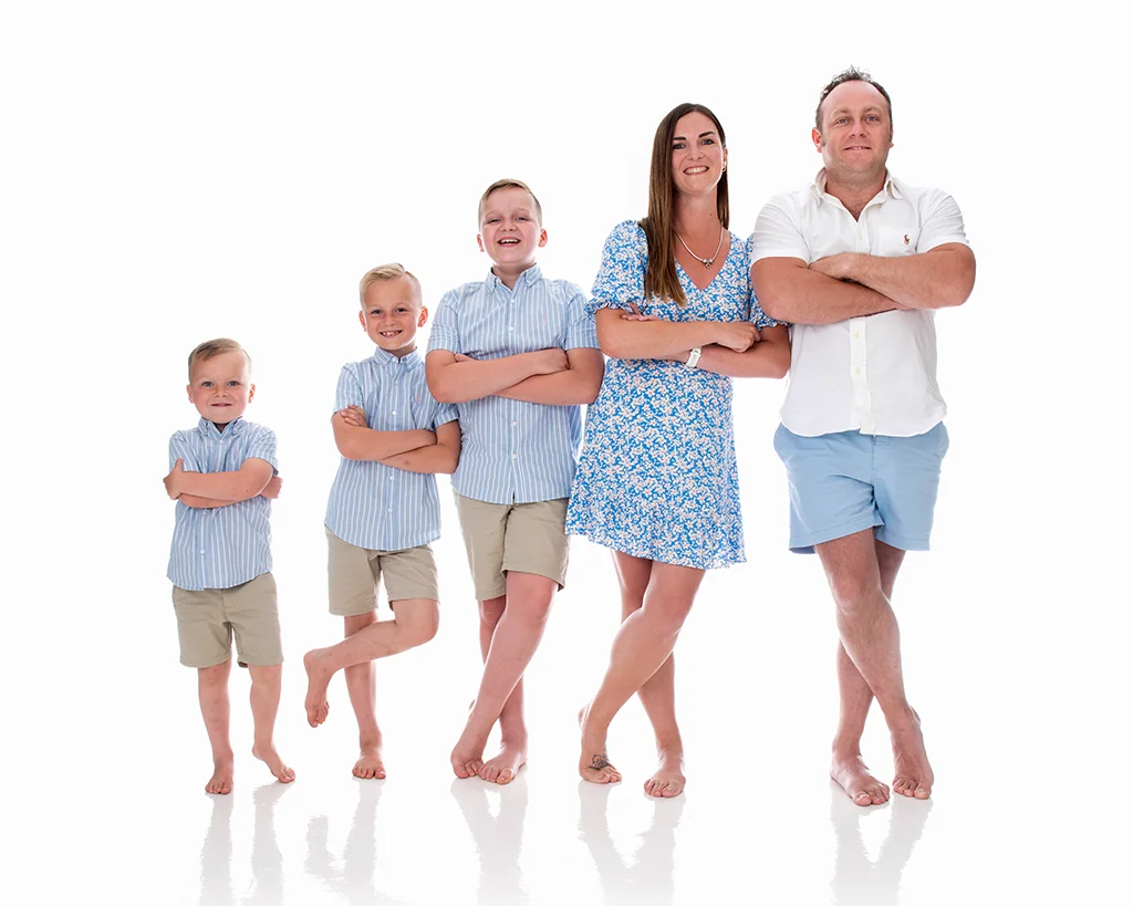 Mum and dad with their three sons stand leading on each other in a completely white room. The boy wear blue shirts and khaki short, the mum a blue summer dress and the dad a white shirt and pale blue shorts