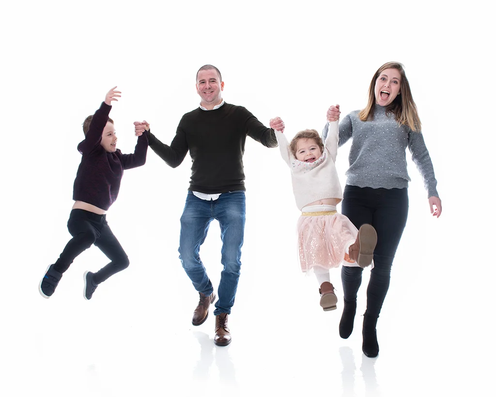 A mum and dad swing their daughter between them while their son holding the dad's hand jumps in the air in a white room photoshoot