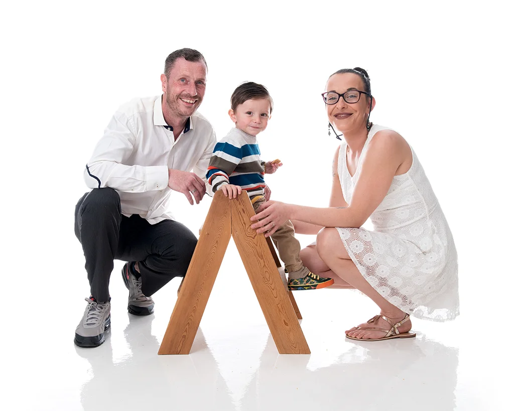 Mum and dad wearing white either side of their son who is sitting on top of a small set of wooden step in a white room photography studio