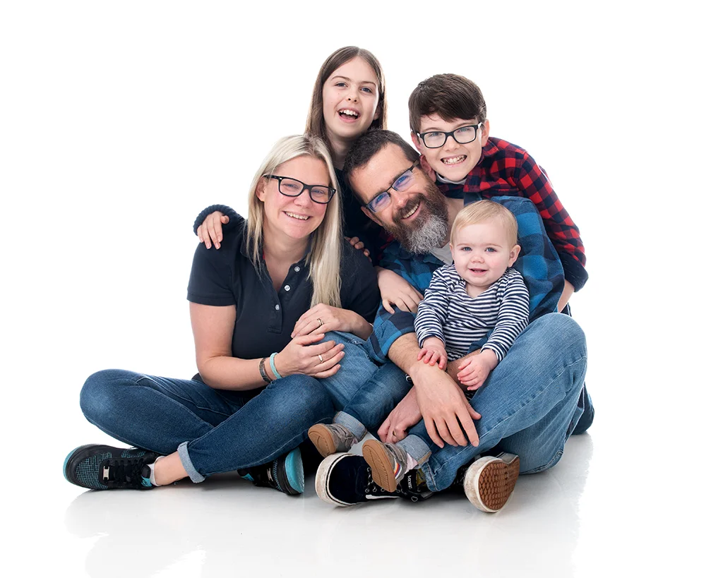 A family of five in a contemporary white room photoshoot. Mum and dad wearing glasses with two older children 10 and 12 with a very young baby girl.