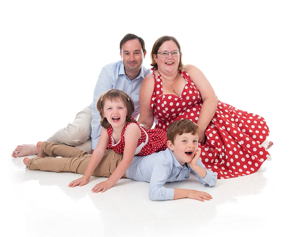 Mum and daughter in bright red dresses with white spots on with her husband and son wearing pale blue Oxford shirt with light colour trousers sitting on the floor in a completely white room
