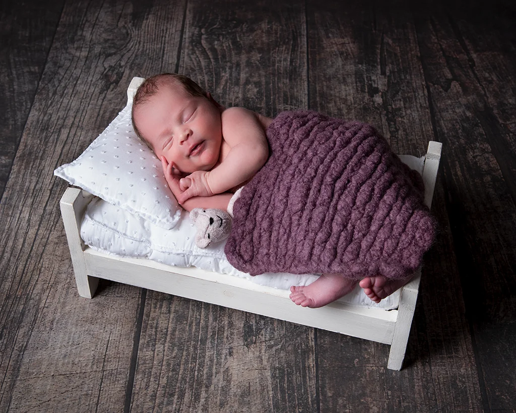 Newborn baby sleeping on a tiny white bed sitting on a brown wood floor with a very small white teddy bear covering in a purple blanket.