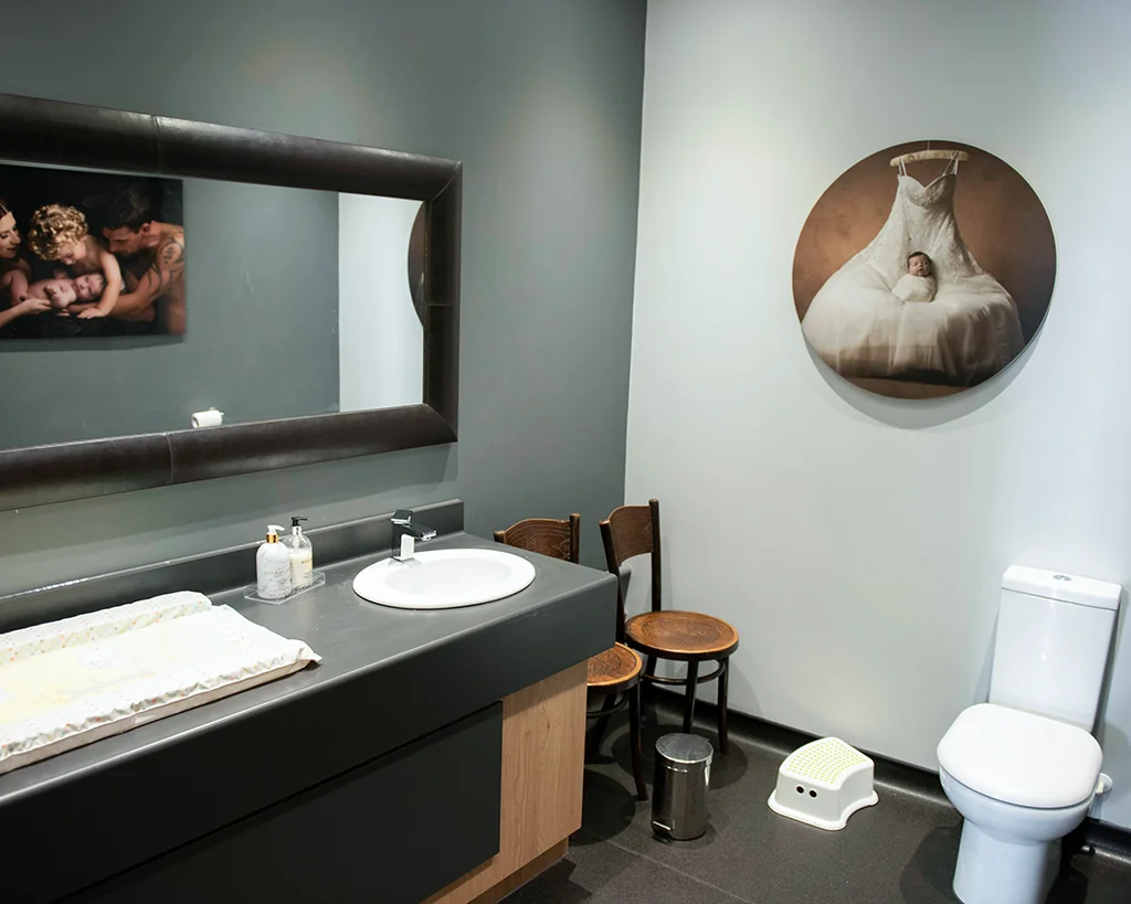 A bathroom with a toilet, sink and baby change table. A loo mirror down the left hand wall and a circular picture of a baby resting on a wedding dress.