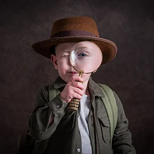 Young boy in an explorer outfit looking through a magnifying glass so he has a big eye