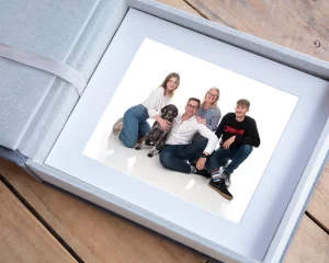 A folio box with 10 family photos mounted prints inside resting on a wood plank surface. The op picture shows a family of four with their dog in a white studio space.