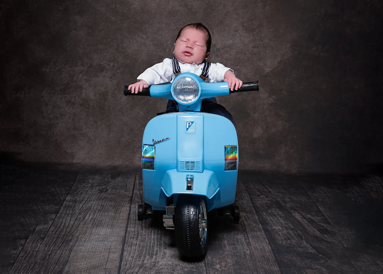 Newborn baby posed asleep on a miniature toy scooter.