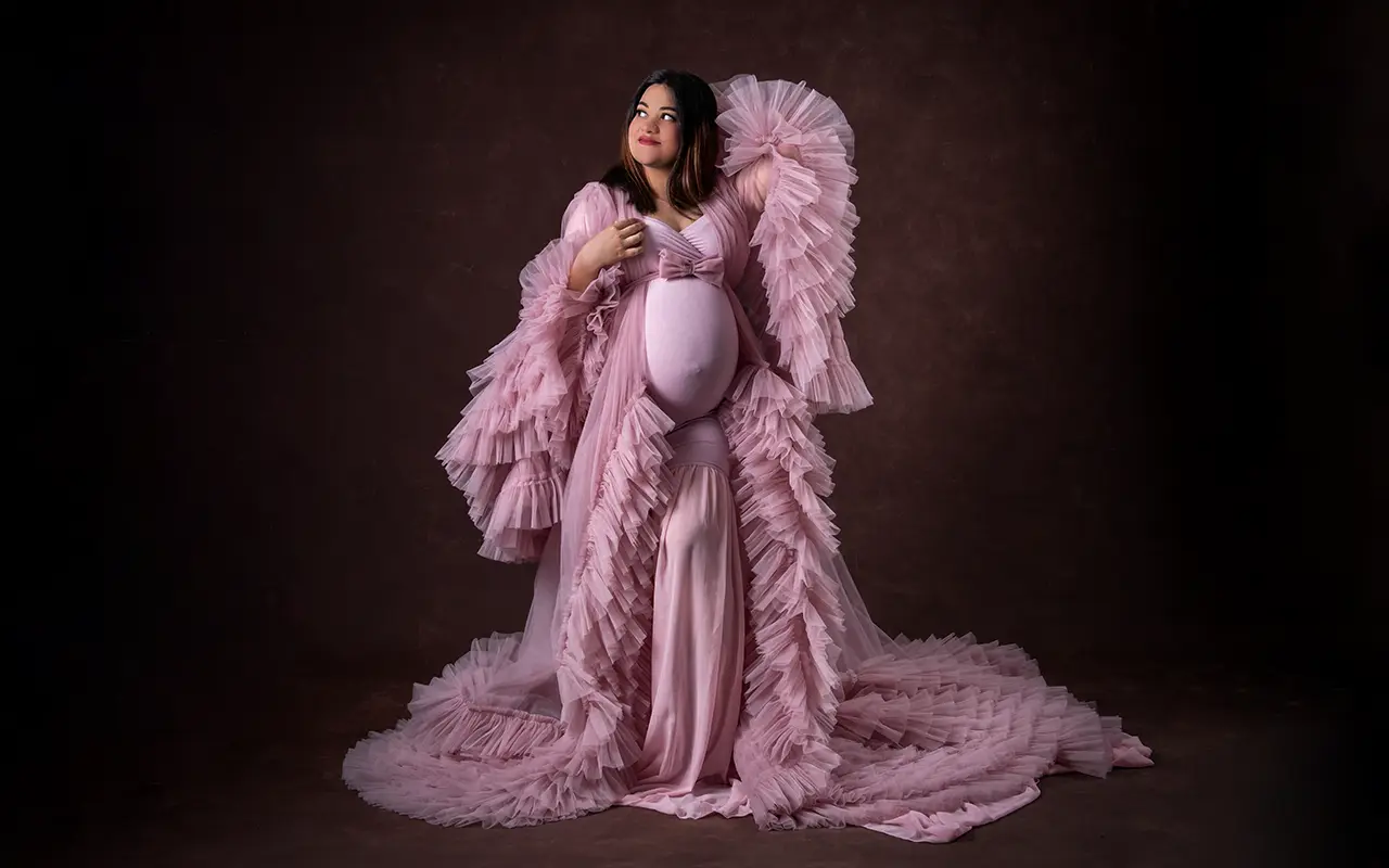 A pregnant woman in an elegant, flowing pink gown poses for a photoshoot.