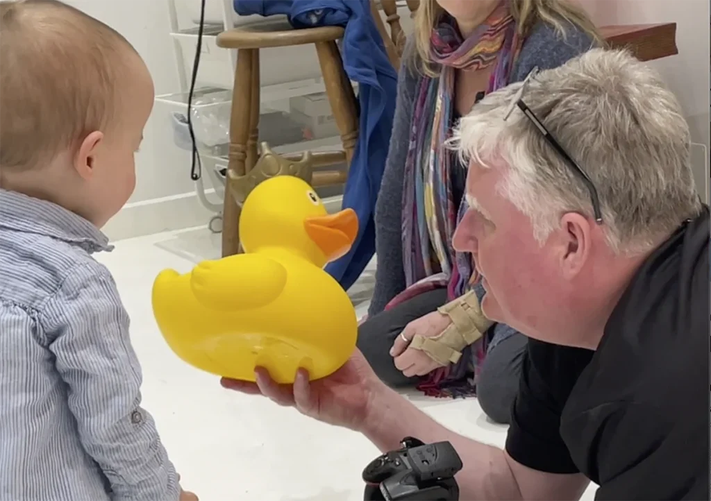 A man showing a large yellow rubber duck to a baby in a room with another adult partially visible.